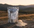 Silver package of energy powder with a scoop on a rocky surface with mountains in the background